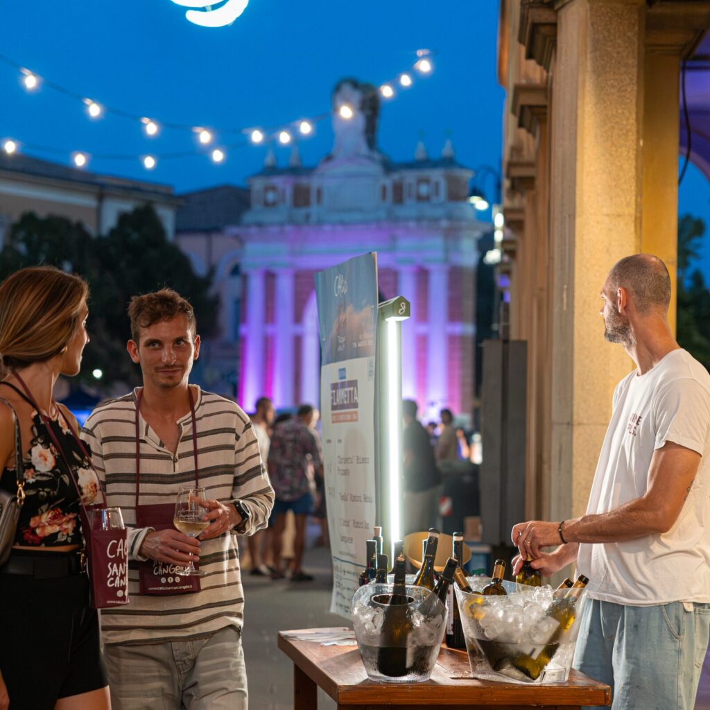 Visitatori con il calice in mano sotto i portici, con la piazza illuminata sullo sfondo
