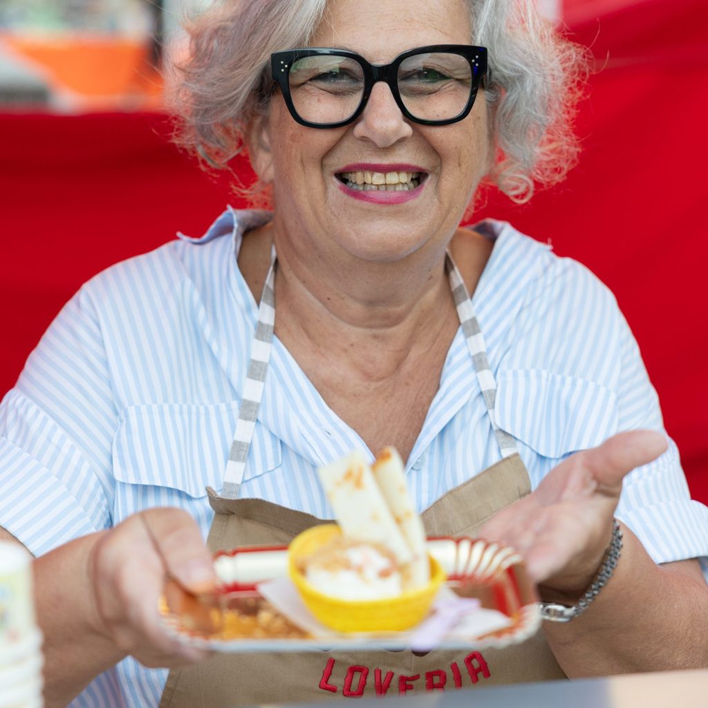 Donna sorridente con grembiule LOVE PIA che porge un piatto di cibo allo stand